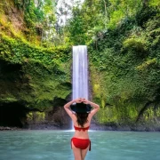 Woman in a red bikini standing in front of a tall, lush green waterfall in Bali, surrounded by dense vegetation