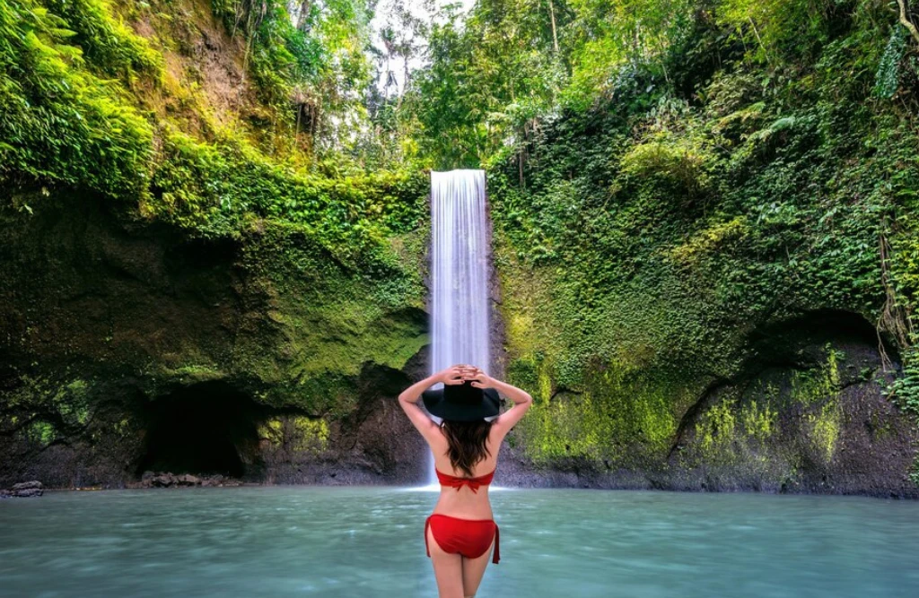Woman in a red bikini standing in front of a tall, lush green waterfall in Bali, surrounded by dense vegetation