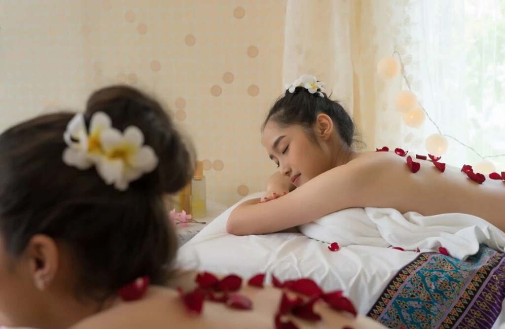 Two women receiving a traditional Balinese massage, lying on massage tables with flower petals, in a calm and relaxing spa environment